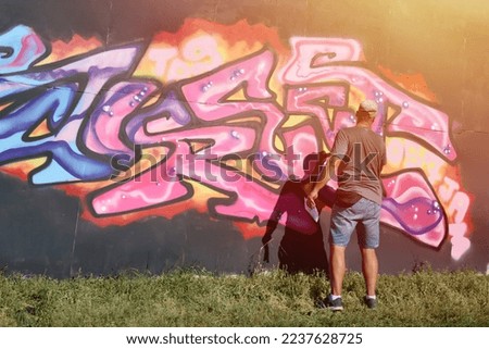 Young caucasian male graffiti artist drawing big street art painting in blue and pink tones. Stylish man in denim shorts and grey t shirt made new graffitti artwork outdoors in bright sunny day Royalty-Free Stock Photo #2237628725