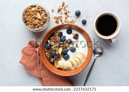 Yogurt with granola, banana, blueberries and dark chocolate chunks in a bowl served with cup of black coffee. Top view healthy breakfast meal Royalty-Free Stock Photo #2237632171