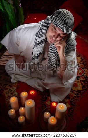 Arab sheikh in a white robe and a headscarf in a boudoir in a room with a red carpet and candles with smoke. fabulous style photo shoot with a man. Male model posing 