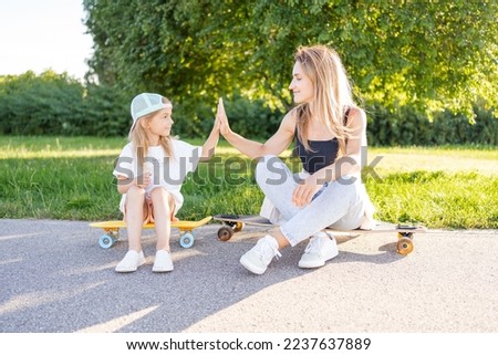 Mother and daughter relax in park sitting on skateboards , give five to each other, spend time together, active family weekend .