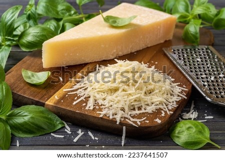 Shredded parmesan cheese and grater on a cutting board. Grana padano cheese whole wedge and grated, stainless steel grater and green basil herb over wooden background. Hard cheese. Front view. Royalty-Free Stock Photo #2237641507