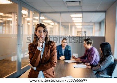 Young happy businesswoman during a meeting in the office looking at camera. Her colleagues are working in the background. Royalty-Free Stock Photo #2237655599