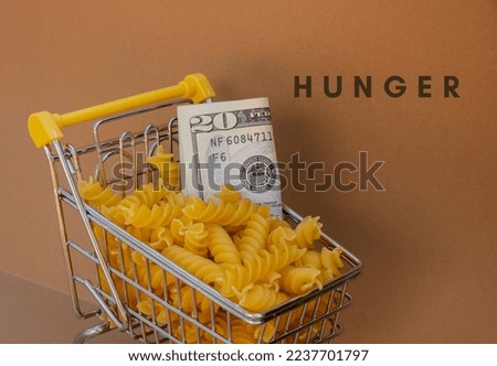 HUNGER text against Shopping trolley cart Filled With Pasta with 20 US dollar paper money banknote on Beige background. Food and groceries shopping price increase, Rising food cost food crisis