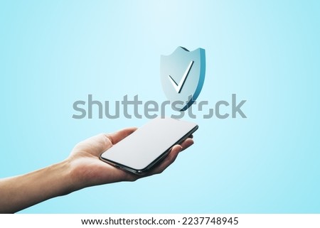 Close up of female hand holding mobile phone with creative antivirus shield icon on light blue background. Secure phone usage, protection and web safety concept