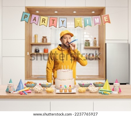 Casual young man preparing for a party and blowing a party horn behind a counter with cake and favors in a kitchen

