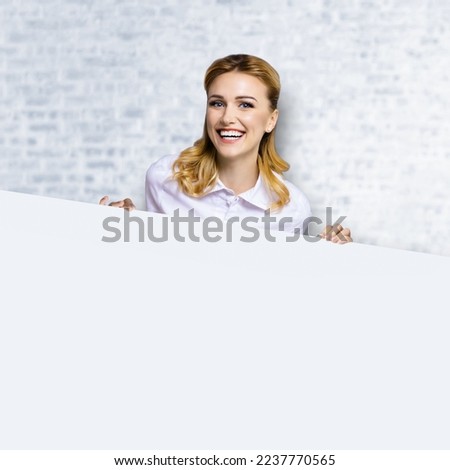 Very happy excited, laughing business woman in white confident clothing standing behind empty banner signboard with copy space area. Success ad concept. White bricks wall. Square image.