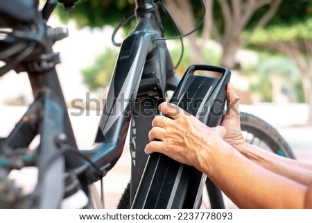 Close up view on senior cyclist woman's hands in urban park fitting the electric battery to his electro bicycle.  Concept of healthy lifestyle and sustainable mobility