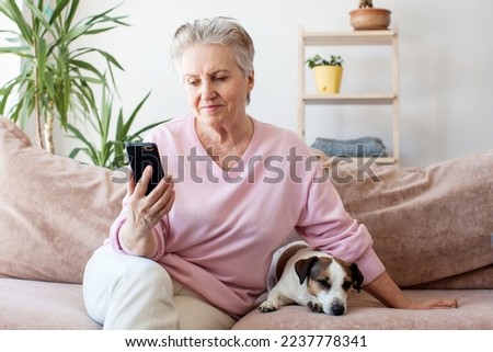 Happy middle aged mature woman enjoying using mobile apps texting typing messages sit on sofa, smiling old adult lady holding smartphone looking at cellphone screen browsing social media at home

