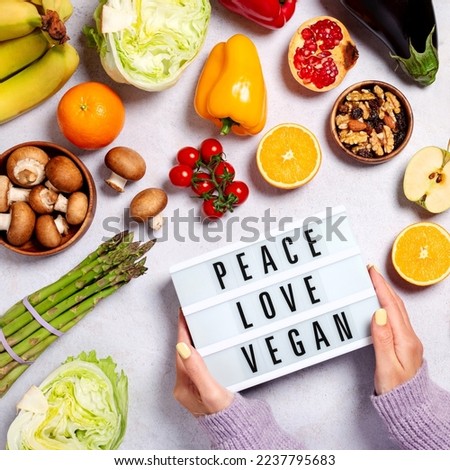 Woman holding lightbox with quote Peace, Love, Vegan on table with raw vegan foods, top view, veganism concept. Raw fresh vegetables and fruits on table with text, vegan food on table