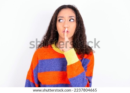 teen girl with curly hair wearing striped knitted sweater over white background makes silence gesture, keeps index finger to lips makes hush sign. Asks not to share secret.
