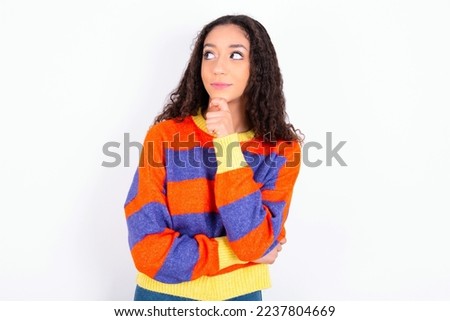 Thoughtful teen girl with curly hair wearing striped knitted sweater over white background holds chin and looks away pensively makes up great plan