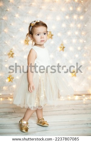 Portrait of a cute baby girl in a white and gold dress and small shiny shoes over Golden Sparkly Background with stars and lights. Magical Christmas time.