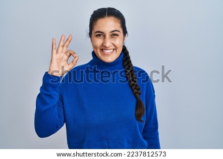 Young hispanic woman standing over isolated background smiling positive doing ok sign with hand and fingers. successful expression. 