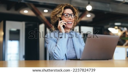 Business woman talking on a phone call in a coworking office. Happy female professional calling her associates on a mobile phone. Professional business woman sitting with a laptop.