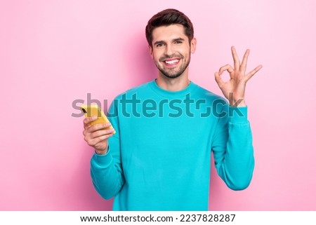Photo of young smiling satisfied brunet hair businessman wear blue sweater show okey sign smartphone recommend shop isolated on pink color background