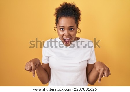 Young hispanic woman with curly hair standing over yellow background pointing down with fingers showing advertisement, surprised face and open mouth 