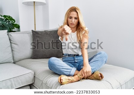 Beautiful blonde woman sitting on the sofa at home looking unhappy and angry showing rejection and negative with thumbs down gesture. bad expression. 