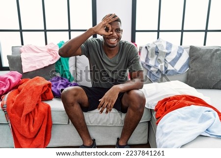 Young african american man sitting on the sofa with dirty laundry clothes doing ok gesture with hand smiling, eye looking through fingers with happy face. 