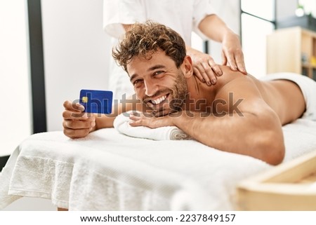 Young hispanic man having back massage holding credit card at beauty center