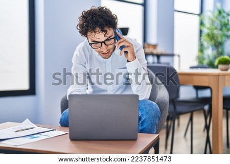 Young hispanic man business worker using laptop talking on smartphone at office
