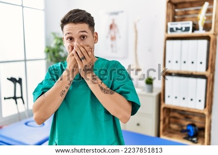 Young physiotherapist man working at pain recovery clinic laughing and embarrassed giggle covering mouth with hands, gossip and scandal concept  Royalty-Free Stock Photo #2237851813