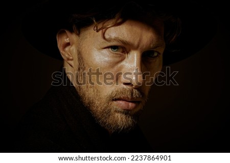 Close up portrait of a pensive mature man with beard and moustache dressed in black coat and black hat looking calmly and thoughtfully. Black background. Psychological picture.