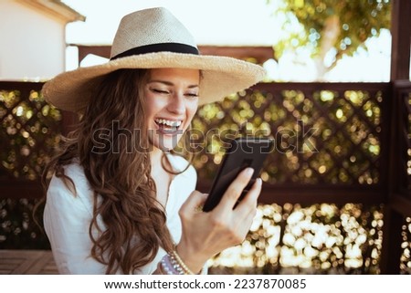 smiling trendy 40 years old housewife in white shirt with hat having video meeting on a smartphone in the terrace of guest house hotel.