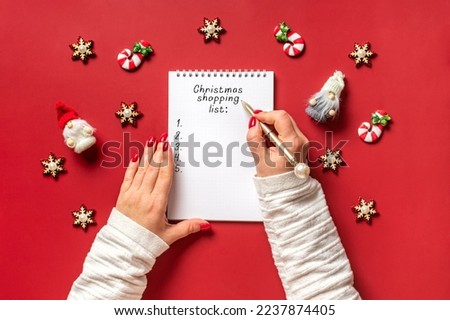 female hands holds pen and writing Christmas shopping list, gift ideas on white notepad on red background Top view Flat lay Holiday card Woman making Christmas presents list, Planning, buying concept Royalty-Free Stock Photo #2237874405