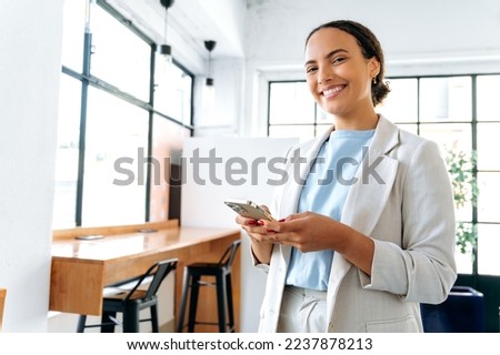 Photo of a positive hispanic or brazilian woman, business entrepreneur, standing in modern office, uses her mobile phone, messaging online with colleague or client, looks at camera, smiling friendly Royalty-Free Stock Photo #2237878213
