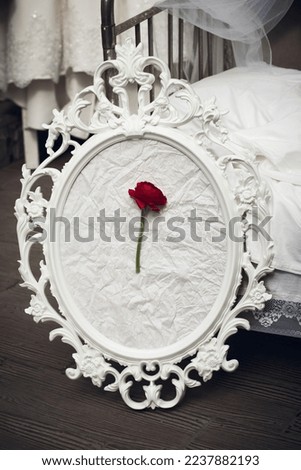 The original composition of a red rose on a white paper background in a vintage frame in a baroque style, stands on a dark wooden floor, in a bright bedroom near the bed, in the morning.
