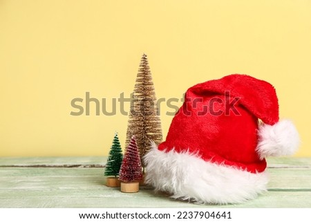 Santa hat and decorative Christmas trees on light wooden table against color background