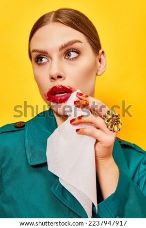 Young woman in green coat with smudged red lipstick wiping mouth after eating nuggets over yellow background. Food pop art photography. Complementary colors. Copy space for ad, text