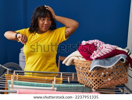 Young beautiful latin woman looking clock hanging clothes on clothesline at laundry room