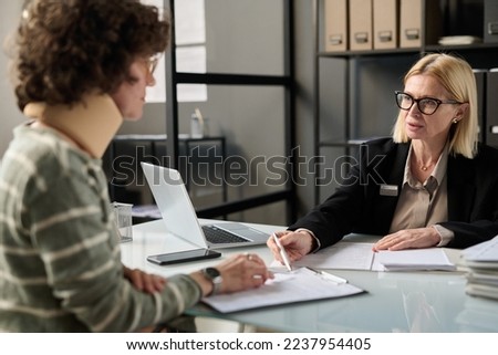 Portrait of female insurance broker consulting injured young woman in office after accident and pointing at legal forms Royalty-Free Stock Photo #2237954405