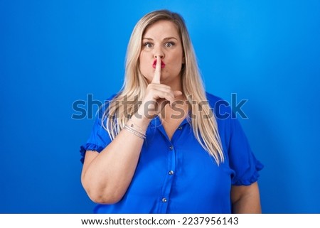Caucasian plus size woman standing over blue background asking to be quiet with finger on lips. silence and secret concept. 