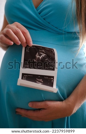 Close-up pregnant woman with ultrasound scan image posing near window at home. Pregnant woman looking at ultrasound scan of baby
