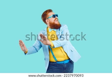 Cool cheerful stylish fat man rejoices and funny dances isolated on light blue background. Emotional red bearded chubby hipster man in good mood dressed in jacket, suspenders, jeans and sunglasses.