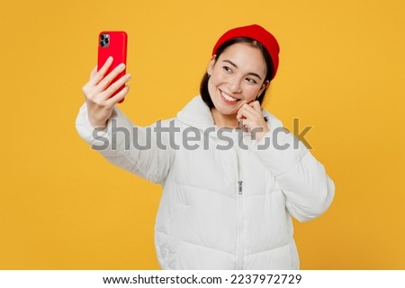 Young happy smiling woman of Asian ethnicity wear white padded windbreaker jacket red hat doing selfie shot on mobile cell phone isolated on plain yellow background studio. People lifestyle concept