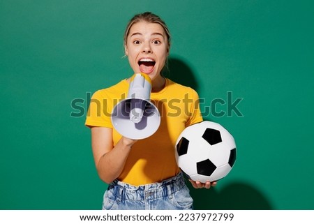 Young woman fan wears basic yellow t-shirt cheer up support football sport team hold in hand soccer ball watch tv live stream hold scream in megaphone announces news isolated on dark green background