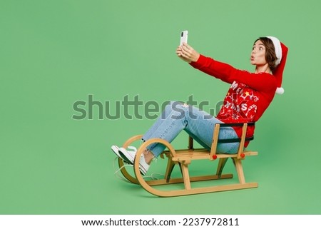 Full size merry young woman wear xmas sweater Santa hat posing sit on sleigh sledding doing selfie shot on mobile cell phone isolated on plain pastel light green background Happy New Year 2023 concept