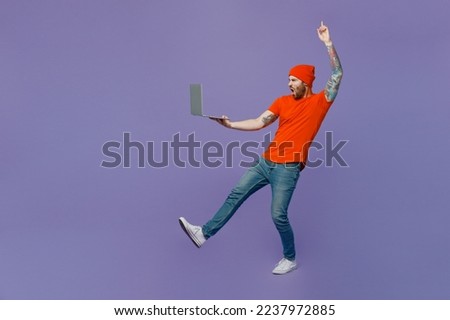 Full body young winner IT man he wear red hat t-shirt hold use work laptop pc computer stand on toes lean back point finger up isolated on plain pastel light purple background People lifestyle concept
