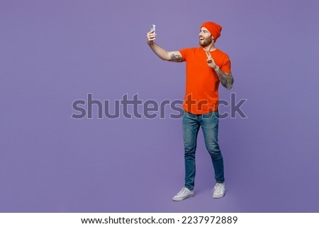 Full body young man 20s he wear red hat t-shirt doing selfie shot on mobile cell phone post photo on social network isolated on plain pastel light purple background studio. People lifestyle concept