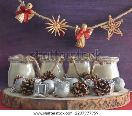 straw christmas decorations hanging on a string under which is an advent wreath with white candles, silver ornaments and gifts, pine cones standing on wood on a purple background