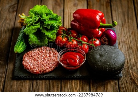 composition of ingredients for beef black burger on a black stone board, raw beef patties, onion, lettuce, tomato, cucumber, ketchup, bell pepper