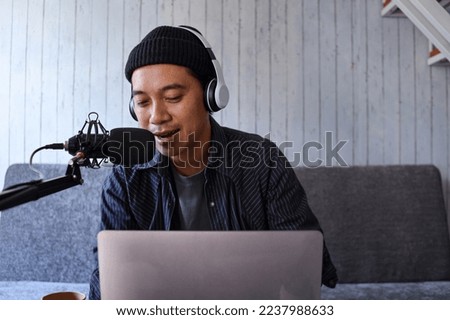 Asian man recording a podcast on his laptop computer with headphones and a microphone at home studio
