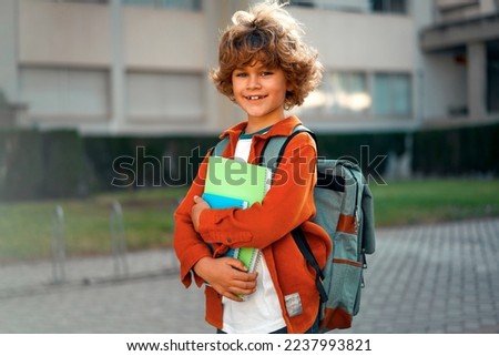 Happy cute smart boy with a stylish hairstyle with a school bag and notebooks in his hand on the street. Modern backpack. First time to school. Back to school.