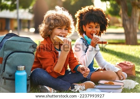 Charming schoolchildren after school resting sitting on the grass in the park. An African-American girl with a Caucasian boy took out lunchboxes with sandwiches from their backpack and eating. Royalty-Free Stock Photo #2237993927