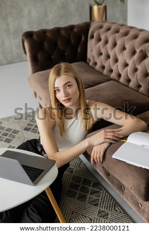 Portrait of blonde coach looking at camera near laptop and notebook at home