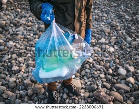 Process of cleaning stone beach from plastic waste. Man holds plastic bottles in trash bag. Environmental pollution concept