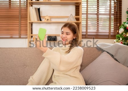 Young asian woman wearing sweater and smiling while sitting to relaxation and using smartphone for taking a photo in holiday on sofa in cozy living room of christmas decoration at home.